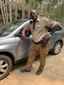Photo: Outdoors an actor stands next to a car.