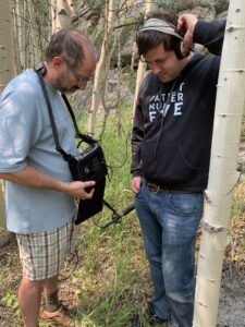 Photo: Outdoors, a man listens through headphones to something another man is playing through a sound recorder.