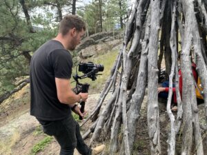 Photo: Outdoors  man holds a camera in front of a wooden tepee.